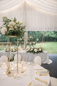 Wesele w plenerze – elegancki stół pod namiotem Elegancki stół weselny ustawiony pod jasnym namiotem w ogrodzie, z jasnymi dekoracjami i złotymi krzesłami.