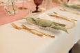 Nakrycie stołu weselnego: złote sztućce, pudrowy róż i oliwkowe dodatki – styl elegancki Zbliżenie eleganckiego nakrycia stołu weselnego: złote sztućce, beżowa serwetka, oliwkowa winietka i pudrowo-różowa aranżacja w tle.